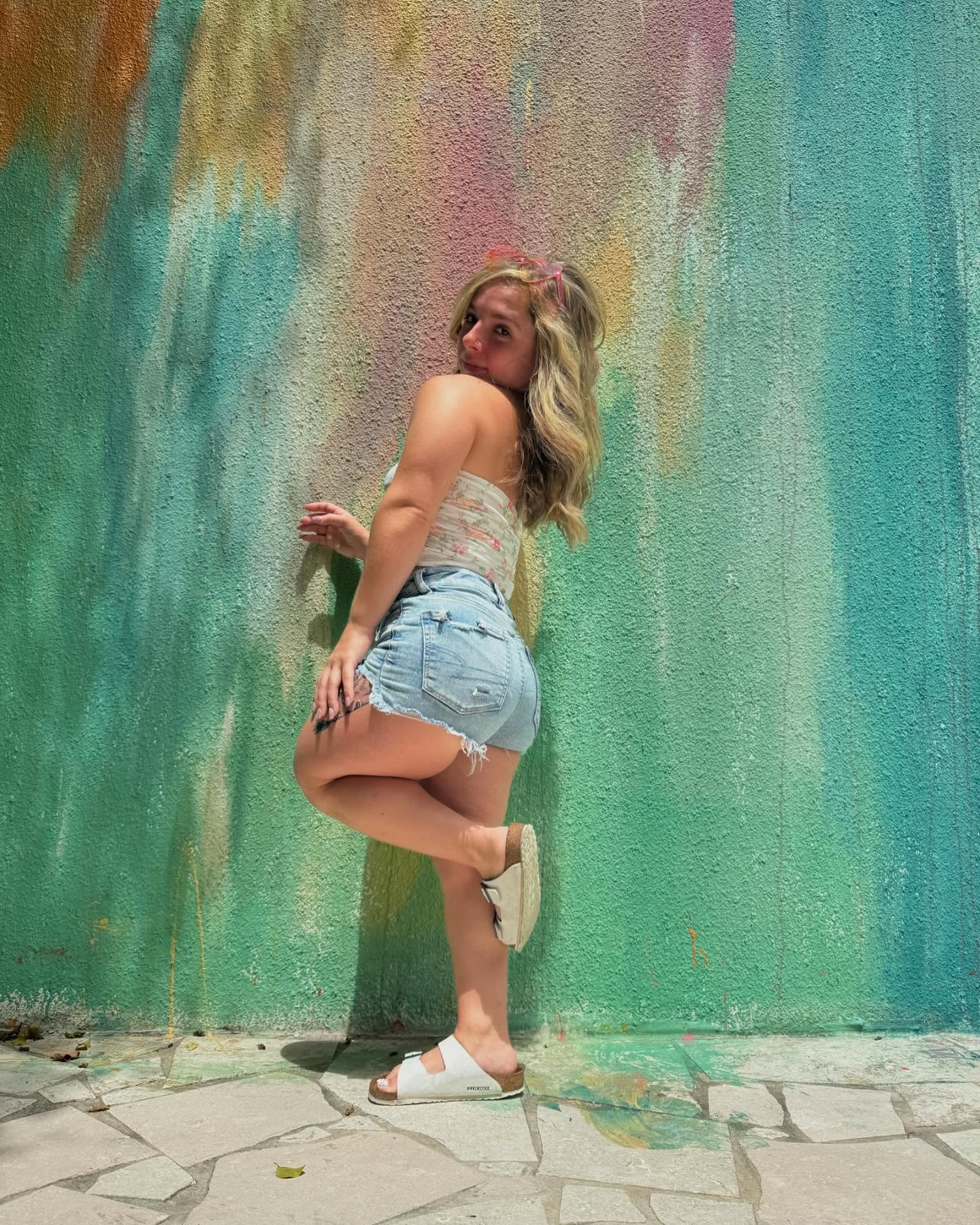 Shy Tayla in denim shorts against a colorful painted wall outdoors